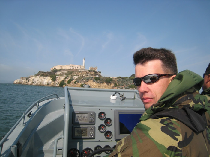 CDR VanVelzen '02 aboard a small vessel in San Francisco Bay, with Alcatraz Island pictured in the background. At the time, VanVelzen was serving in the U.S. Coast Guard. 
